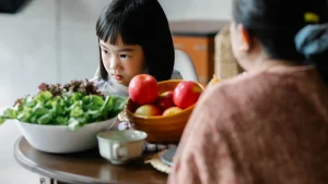 Enfant à table devant un saladier de salade et une corbeille de pommes, avec un adulte