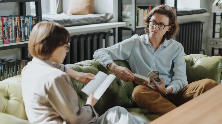 Deux lecteurs assis sur un canapé qui échangent autour d'un livre devant des étagères de livres